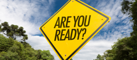 A yellow street sign is shown close up with the words in black "ARE YOU READY?" The background is a blue sky with clouds and trees.