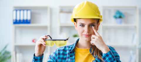 A woman is shown wearing a yellow hard hat, yellow shirt, and blue plaid shirt unbuttoned. She is pointed to her eye with one hand and holding protective safety glasses in the other hand.